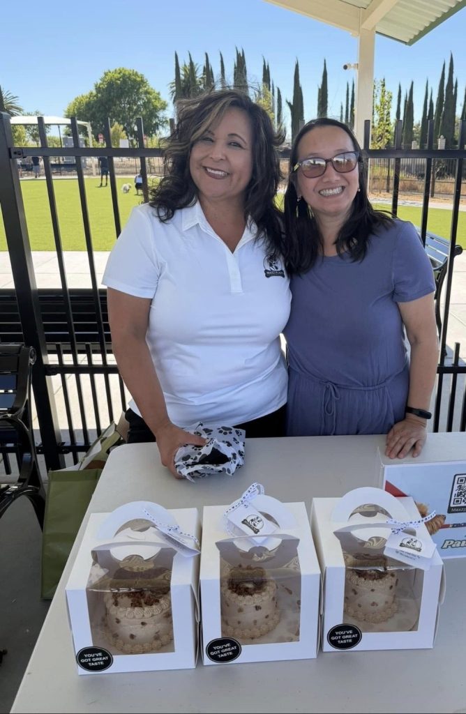 Two women at a table of boxed treats beside the dog park fence